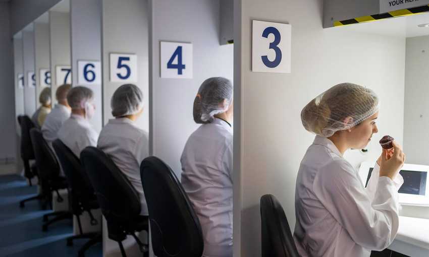 A row of people in white lab coats and hairnets sit at numbered stations. Each person is engaging in a sensory evaluation, with one person smelling or tasting from a small cake under a hooded light. The stations are numbered 3 to 8.