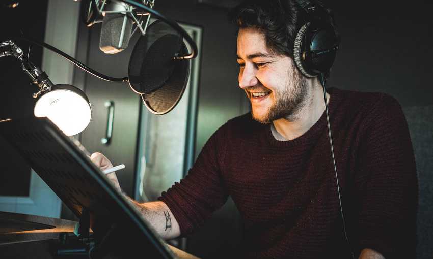 A man wearing headphones smiles while sitting in front of a microphone in a recording studio. He holds a pen and appears to be working or recording audio. The setting is dimly lit, with a lamp illuminating his workspace.
