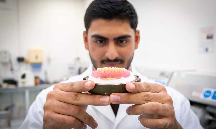 A young man in a white lab coat holds a dental prosthetic.
