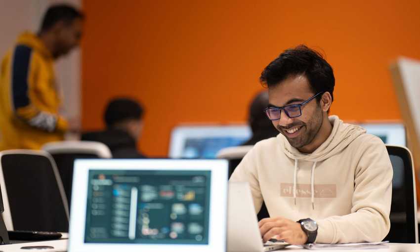 A student wearing a white hoodie and glasses is focused on his laptop while working at a desk.