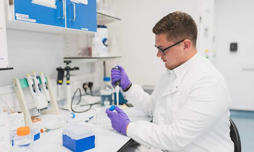 A person in a lab coat uses a pipette to dispense a sample into a container.