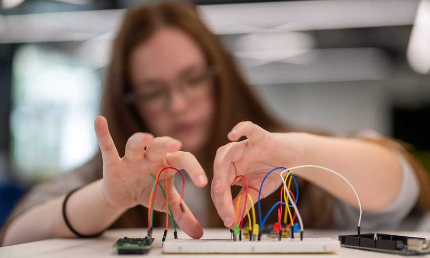 A person with long hair and glasses is working with a breadboard and wires, connecting electronic components. The background is blurred, focusing on the hands and the breadboard.
