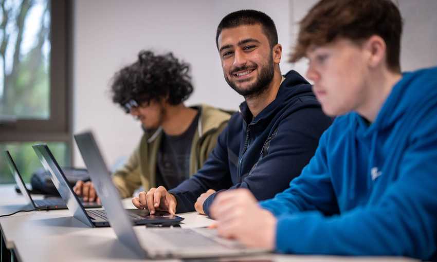 Three people sit alongside each other at a row of desks. Each has a laptop computer in front of them.