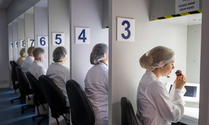 A row of people in white lab coats and hairnets sit at numbered stations. Each person is engaging in a sensory evaluation, with one person smelling or tasting from a small cup under a hooded light. The stations are numbered 3 to 8.