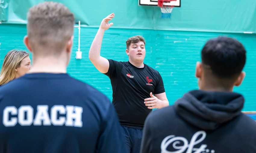 A person stands in a gym, gesturing with one hand raised, facing a group. The back of one person is visible, wearing a shirt labeled Coach. A basketball hoop is in the background. The setting is casual and suggests a team gathering or discussion.