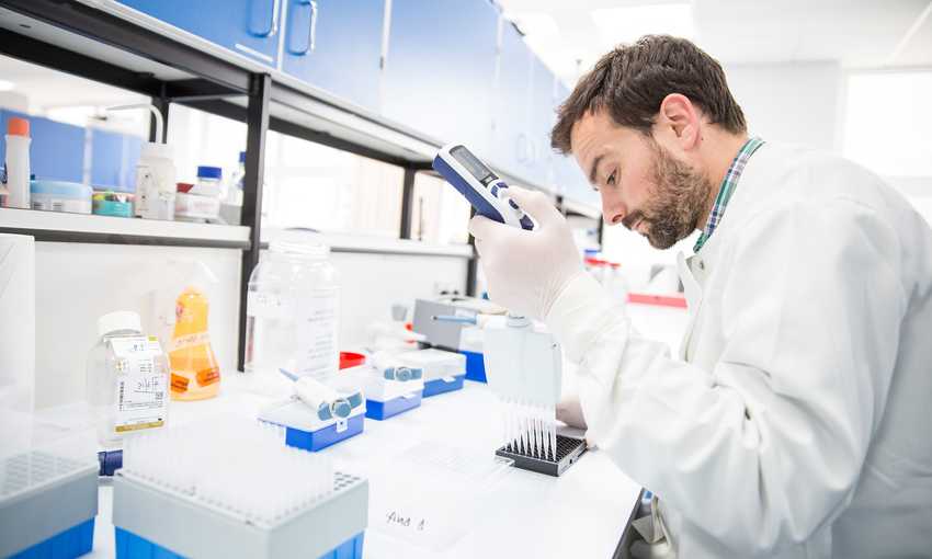 A scientist in a lab coat uses a pipette to transfer liquid into a tray, surrounded by lab equipment and bottles.