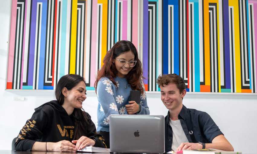 Two people sit at a table in front of a laptop. Behind them stands a third person.