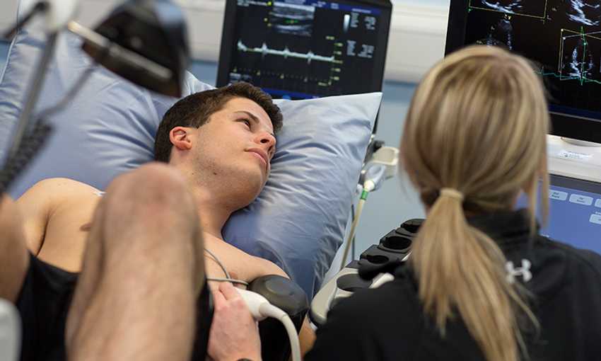 A student undergoes an ultrasound assessment while a technician monitors the results on multiple screens.