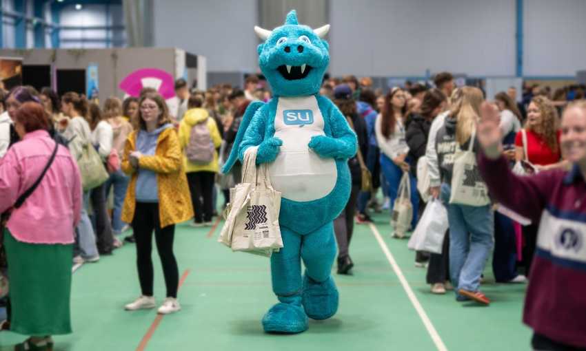 The Students' Union mascot, a light blue and white dragon, holding a tote bag in the middle of a busy Freshers' Fayre