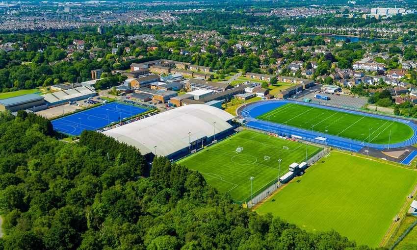 Aerial view of a sports complex featuring a large indoor sports facility, blue athletics track, multiple soccer fields, lush green trees in the foreground, and nearby residential buildings in the background.