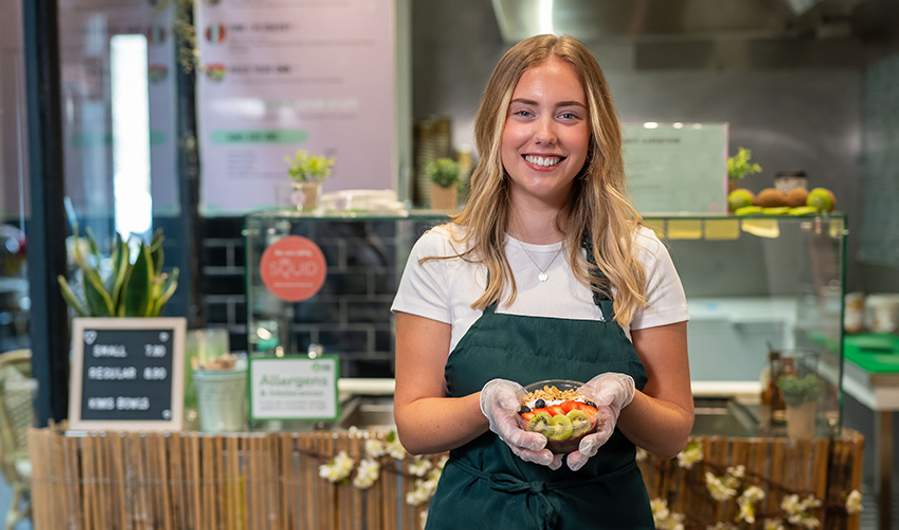 A young person wearing a green apron holds a fruit bowl while standing in front of a kitchen counter