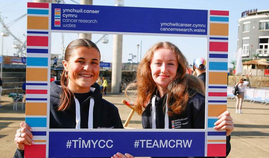 Two people hold a sign for Cancer Research Wales.