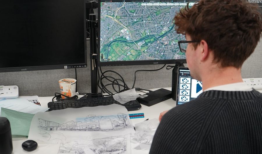 A young man analyses a computer screen and accompanying architecture plans at a desk