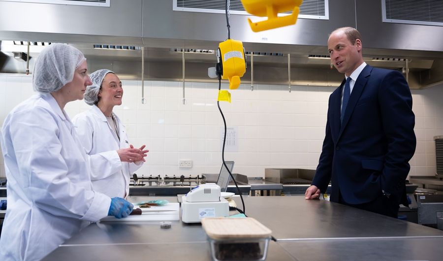 Prince William engages in conversation with two workers in lab coats and hairnets