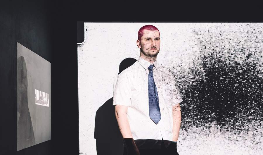 A young man in white shirt and tie stands in the light of a projector screen
