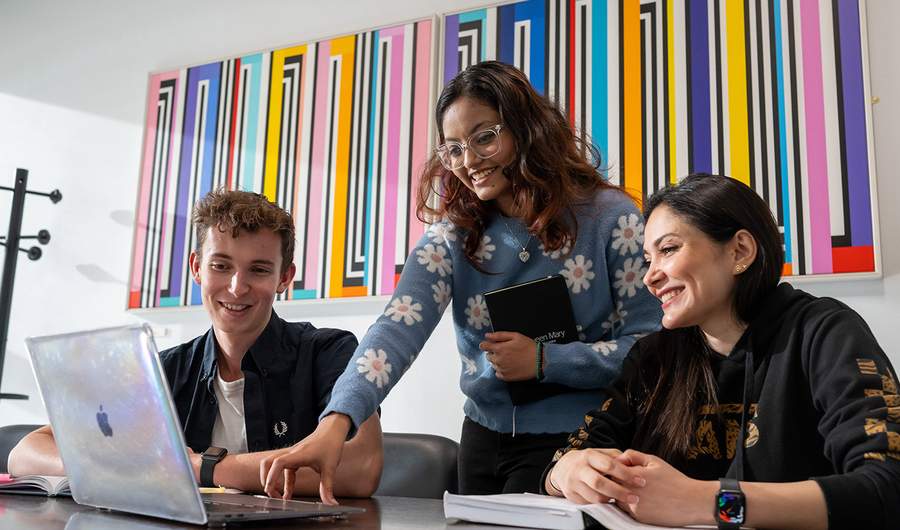 Three students collaborating on a laptop in a modern office setting, focused on their project.