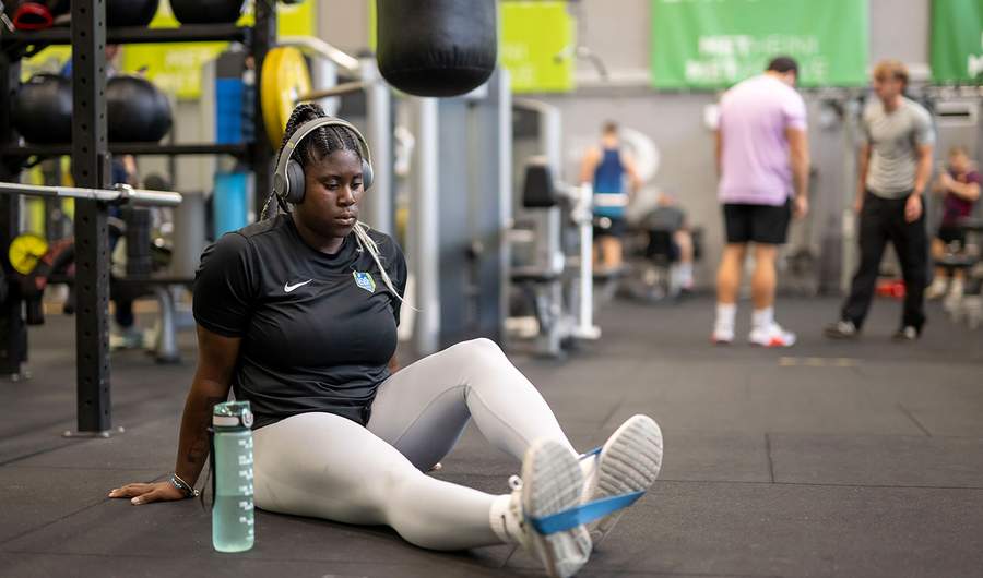 A woman sitting on the gym floor, engaged in a fitness activity, with weights and mats visible around her.