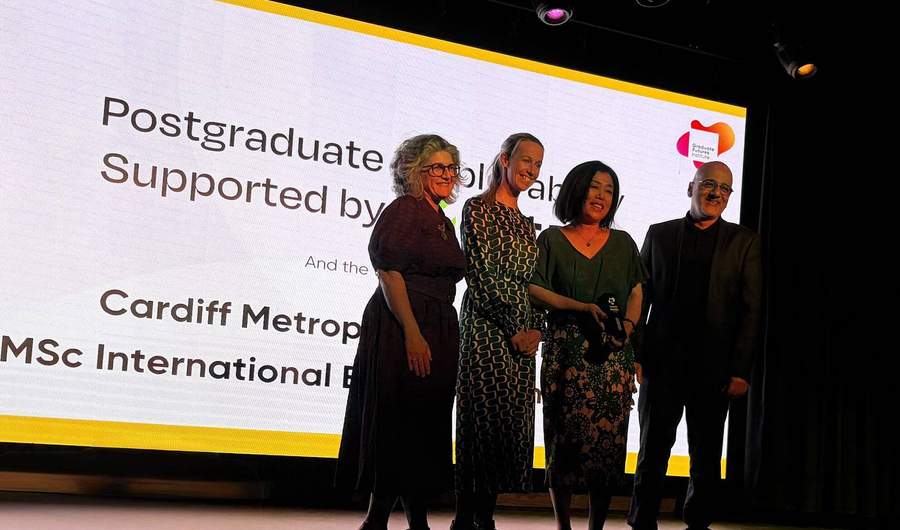 A group of four people pose for a photo in front of large display screen. One of them is holding an award in their hands.