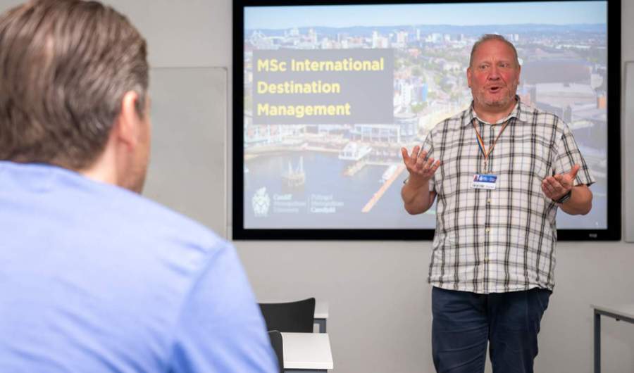 A man stands in front of a screen giving a presentation. The screen shows a view of Cardiff Bay with the caption MSc International Destination Management.