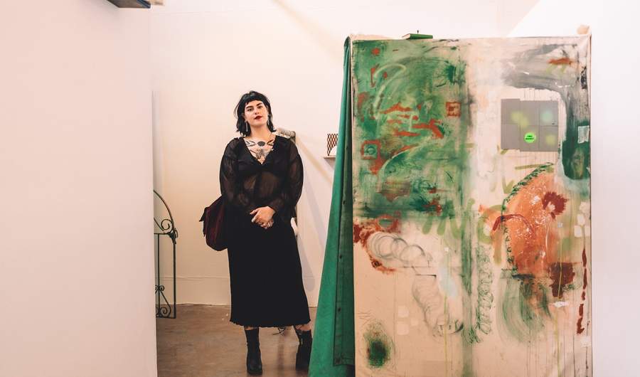 A young woman in a black dress stands beside a large painted canvas