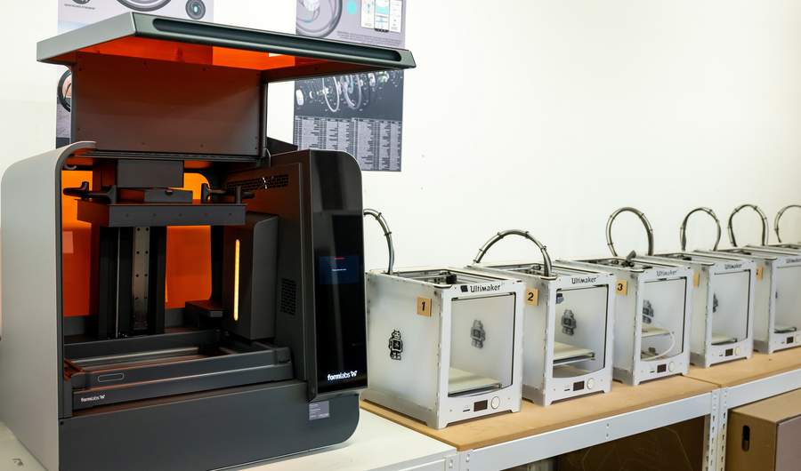 A collection of 3D Printers lined up on a row of desks