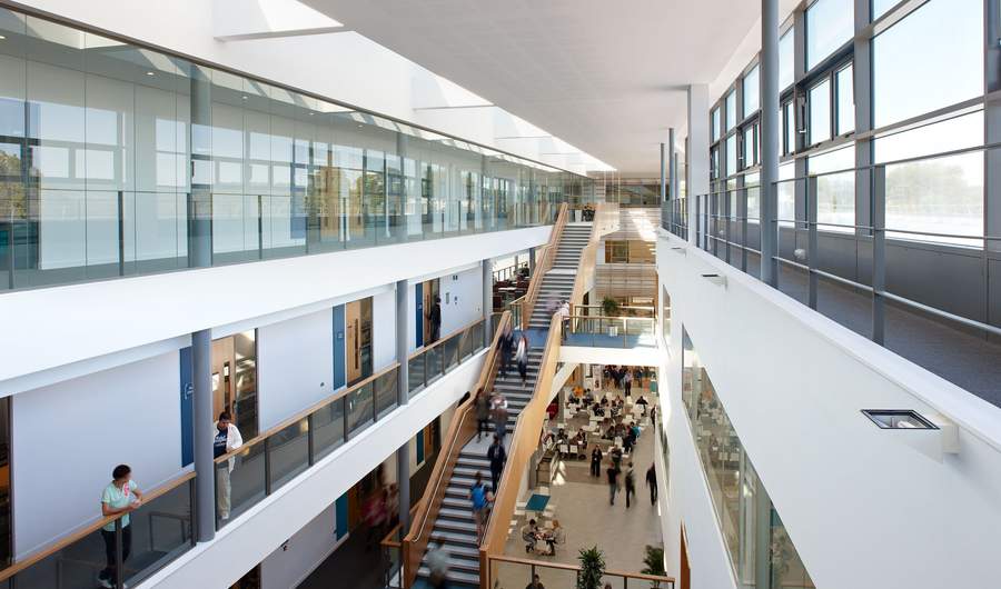 A modern, spacious atrium with a large wooden staircase and multiple people walking. The area has glass railings, plants, and abundant natural light from skylights above. The scene conveys a busy, contemporary office or educational environment.