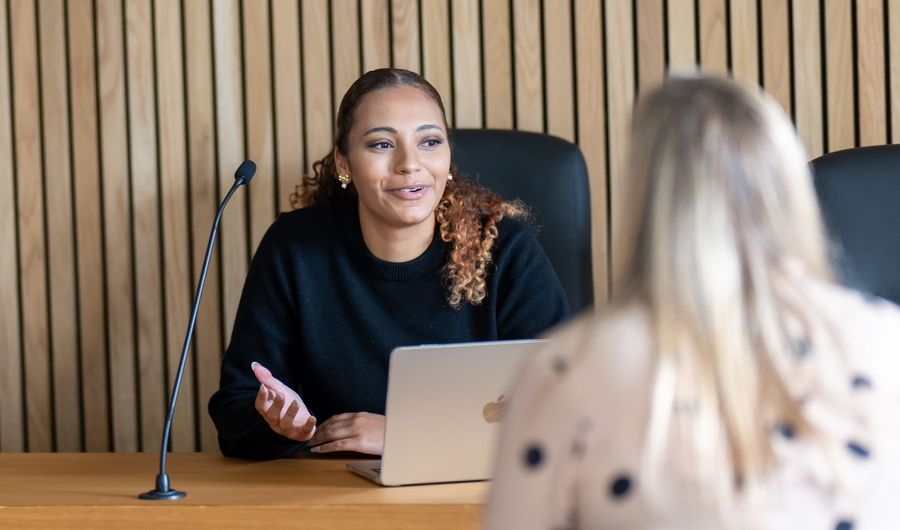 A person sits at a courtroom bench. They are in conversation with someone sat across from them.