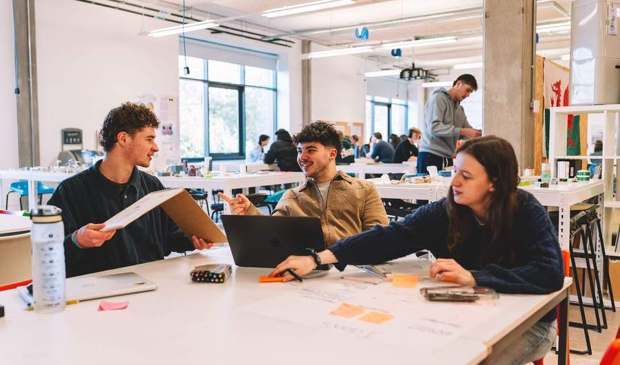 Three students sit together at a table in a studio space. One is holding a sketchbook, and there are laptops, pens and paper on the table.