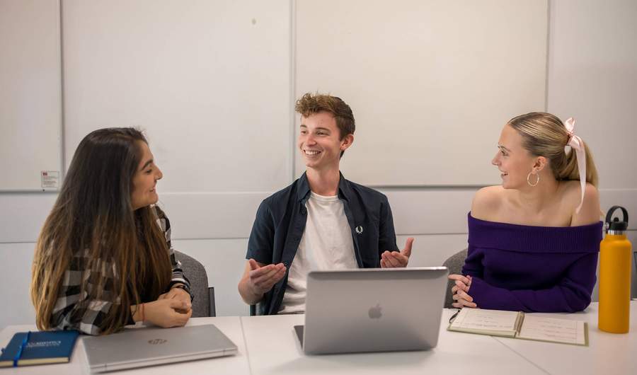 Three students sit alongside each other at a table with laptop computers and notebooks.