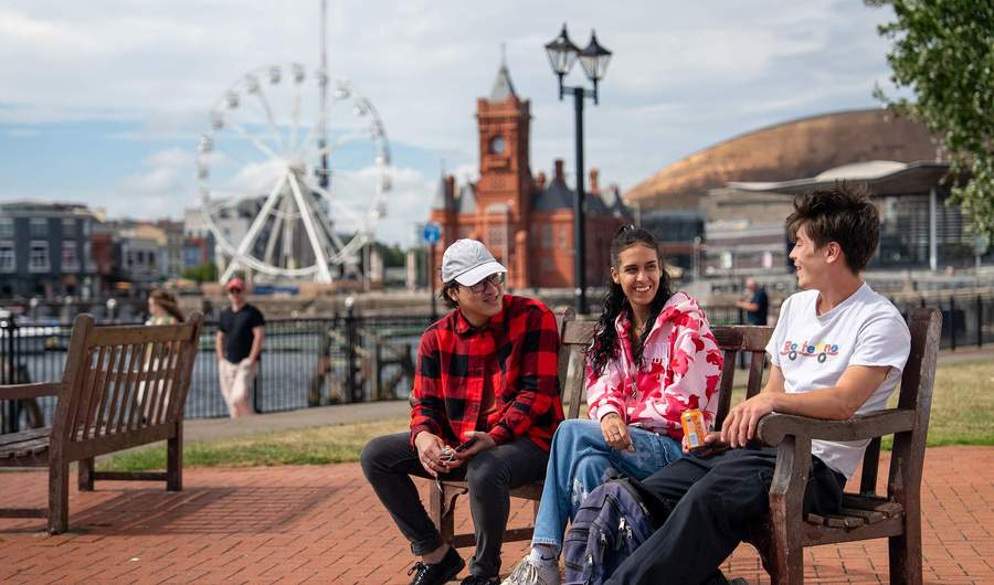 Three students sit on a bench in Cardiff Bay. Behind them is a Ferris wheel, the Pierhead Building and Wales Millennium Centre.