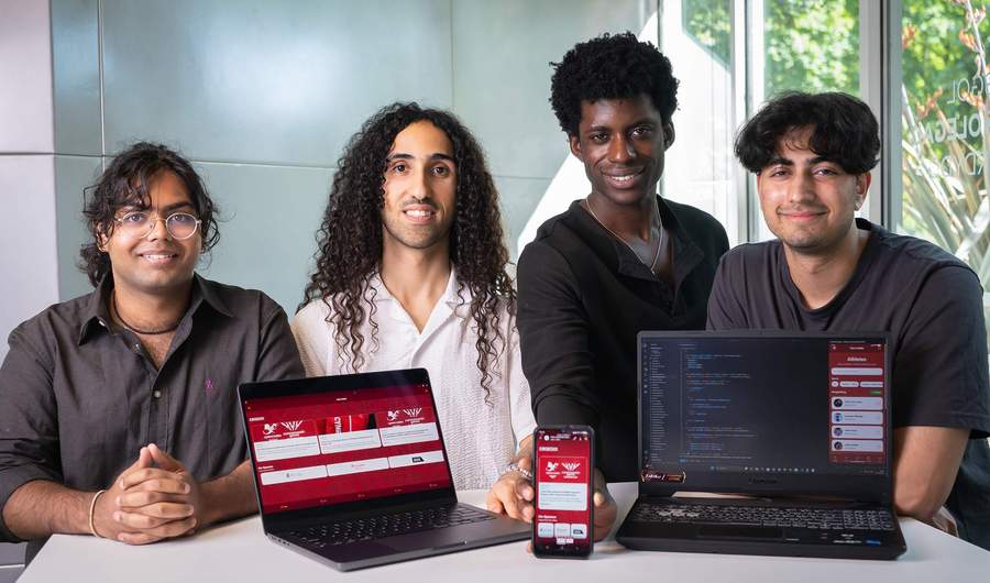 Four men smile for a photo. In front of them are two laptops showing an app in development. One is holding a mobile phone showing the same app.