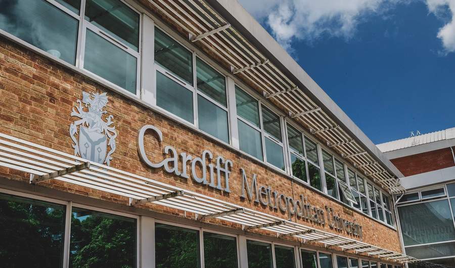 A building with large windows featuring the sign Cardiff Metropolitan University on a brick wall. A blue sky with clouds is visible above.