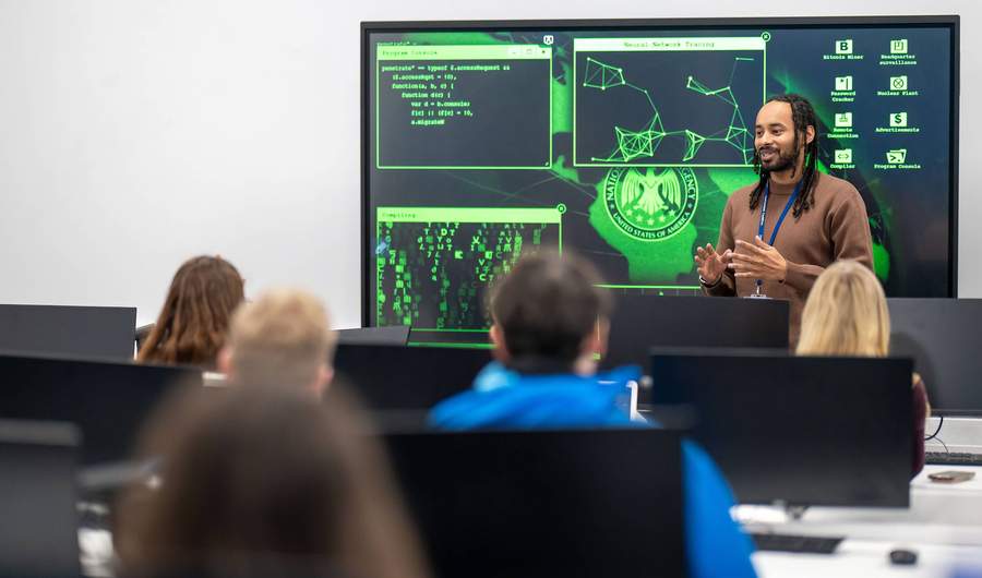 A lecturer stands in front of a large screen displaying computer graphics, engaged in a presentation or discussion.