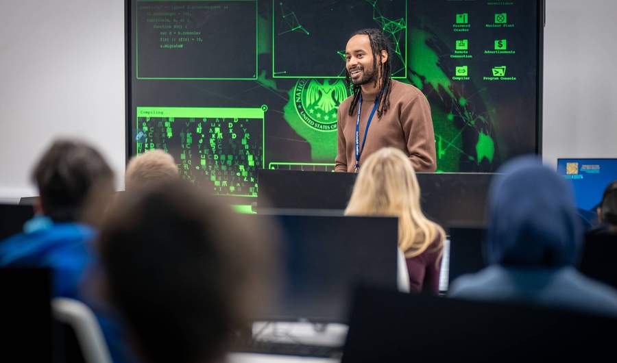 A lecturer stands in front of a large screen displaying computer graphics, engaged in a presentation or discussion.