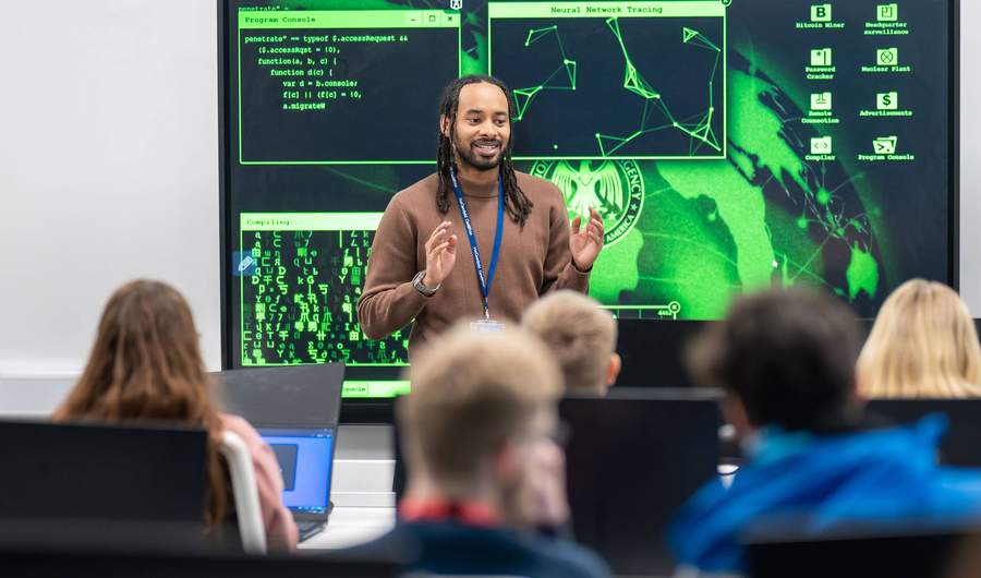 A lecturer stands in front of a large screen displaying computer graphics, engaged in a presentation or discussion.