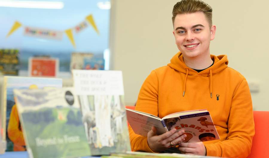 A person in an orange hoodie is smiling while holding a book in a library. There are several books displayed in the foreground. The background shows blurred decor and furniture.