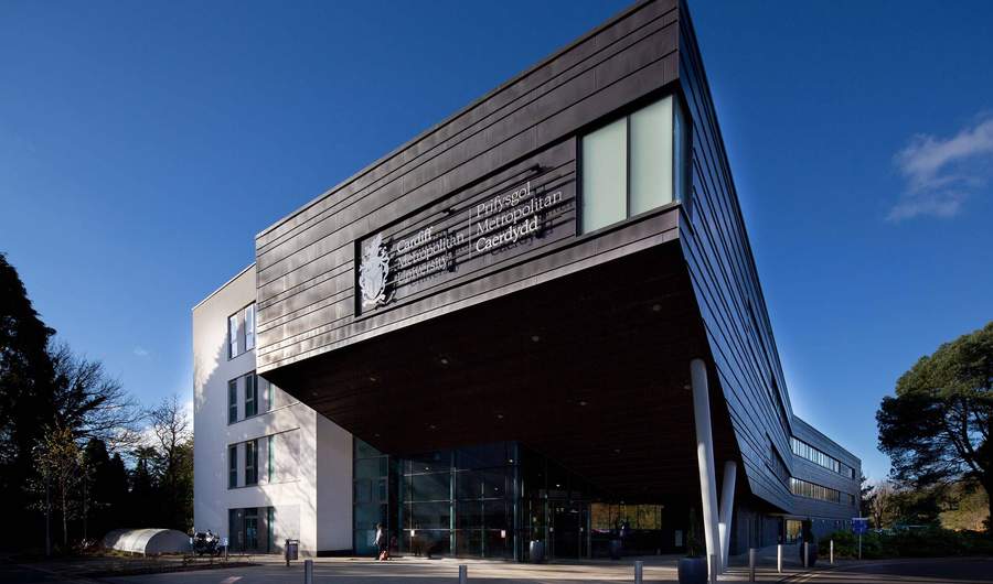 Modern building with angular architecture, featuring a large sign that reads Prifysgol Metropolitan Caerdydd. The structure has a mix of dark panels and white walls, with glass windows. Trees and a blue sky are visible in the background.