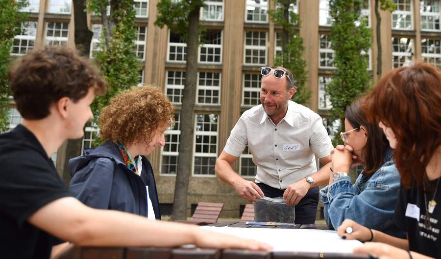 Four people sit at a wooden table outdoors. A man wearing a name badge labelled Gary stands next to them.