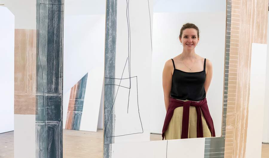 A woman stands smiling amidst an abstract art installation with geometric designs on large panels. She is wearing a black top and has a sweater tied around her waist. The setting appears to be an art gallery.