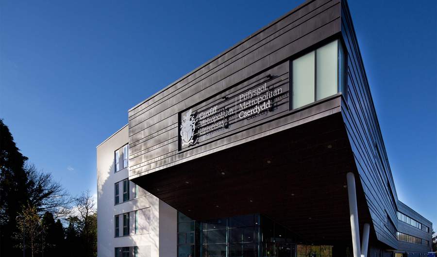 Modern building with angular architecture, featuring a large sign that reads Prifysgol Metropolitan Caerdydd. The structure has a mix of dark panels and white walls, with glass windows. Trees and a blue sky are visible in the background.