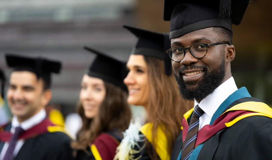 A graduate in cap and gown. In the background are three other graduates also wearing caps and gowns.