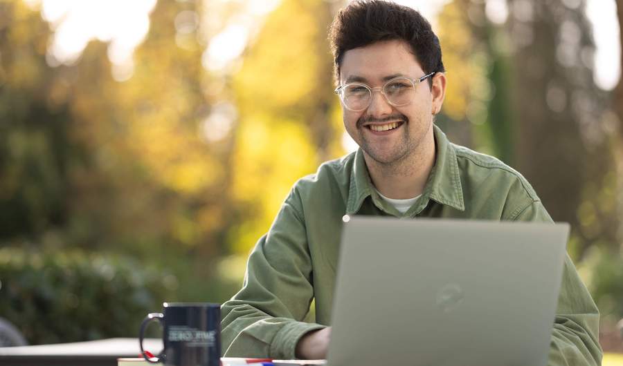 A young man sitting outside at a table. In front of him is a laptop computer and coffee mug.