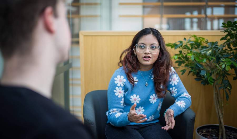 A young woman with glasses and long hair, wearing a blue sweater with white flower patterns, gestures while talking. She is seated in a modern indoor space, facing another person whose back is to the camera. A green plant is in the background.