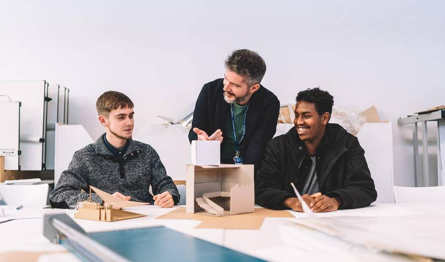 A lecturer stands between two students seated at a table with small architectural models made from card.