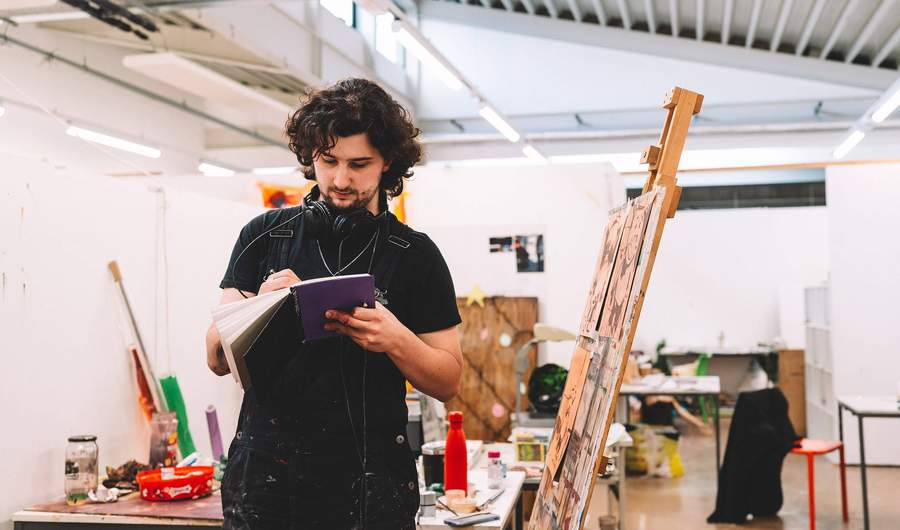 A young man standing next to an easel writes in a notebook.