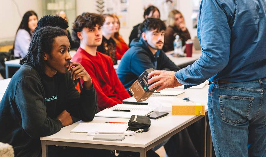 Student being shown a boxed product while seated at a classroom table with others listening nearby.
