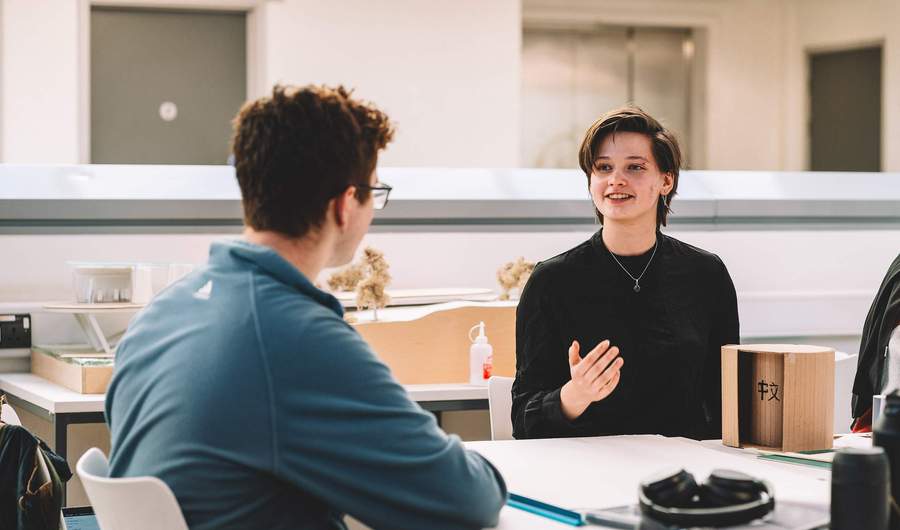 Two students seated at a table discussing a small cardboard model with tools and materials nearby.