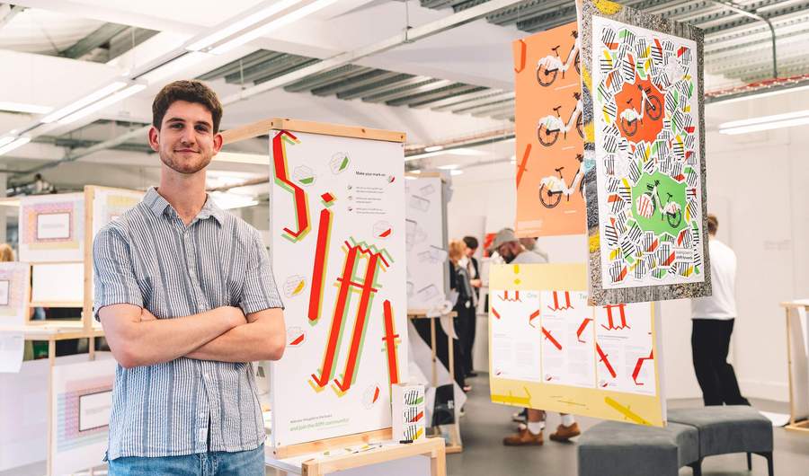 A student stands with arms folded next to a display of design work.