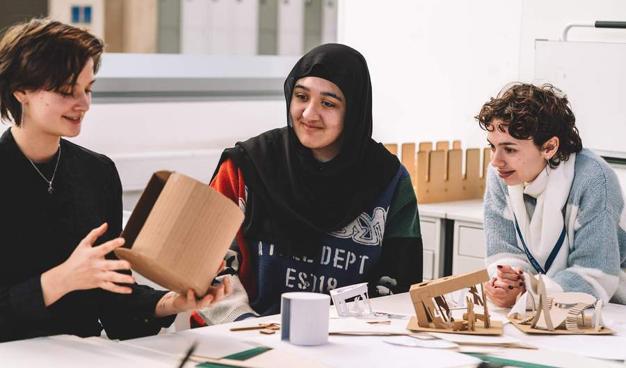 Three students seated at a table reviewing small architectural models and cardboard structures.
