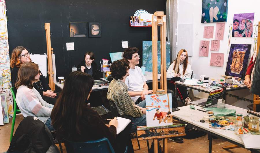 A group of students in a studio space, surrounded by canvas paintings and sketches.